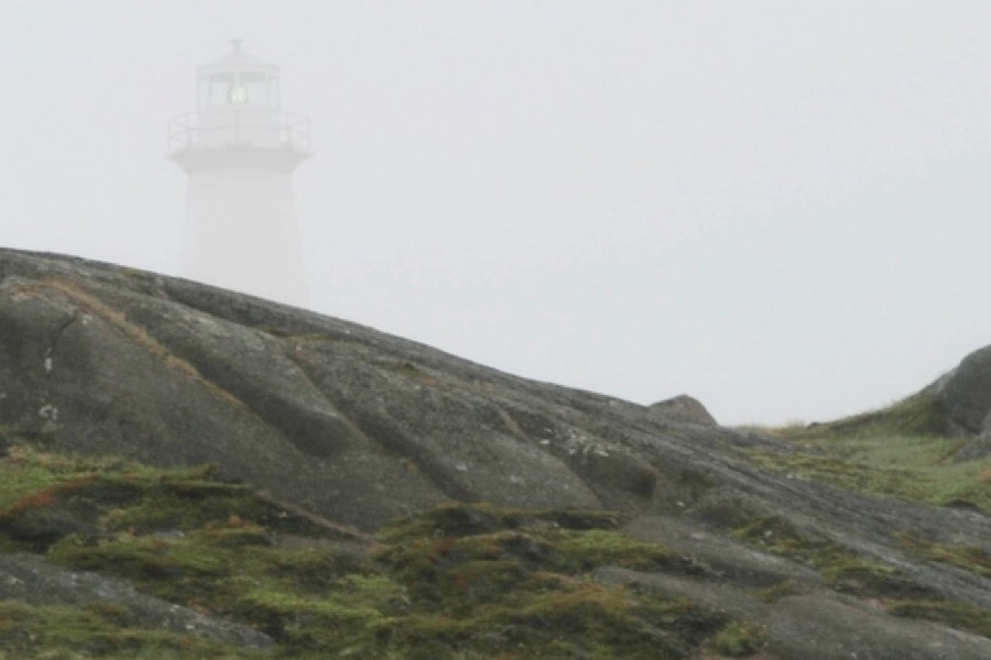 A white lighthouse half hidden in sea fog above grey rocks and moss